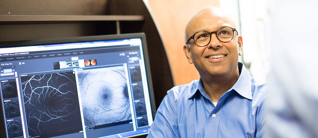 Dr. Gupta smiling with eye scans on screen behind him.